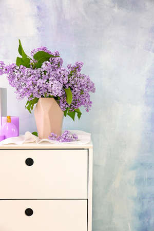 Beautiful lilac flowers in vase on chest of drawers against light background