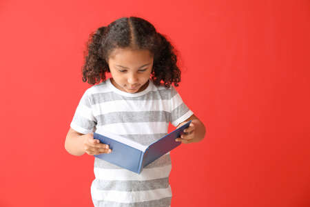 Portrait of adorable little African-American girl reading book on color background