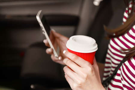 Young woman with mobile phone and coffee in car, closeupの写真素材