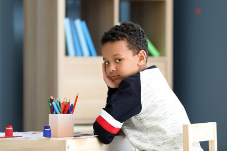 Cute African-American boy sitting at table in room