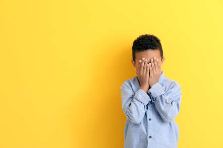 Cute African-American boy covering face with his hands on color background