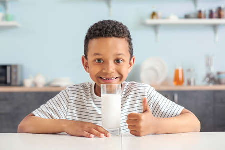 Cute African-American boy with glass of milk showing thumb-up at tableの写真素材