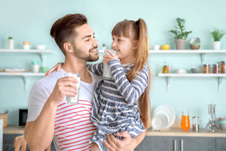 Young father with little daughter drinking tasty milk in kitchen at homeの写真素材
