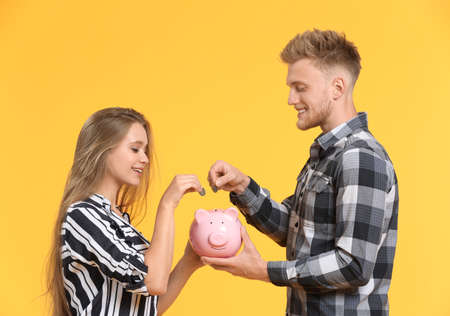 Happy couple putting coins into piggy bank on color backgroundの写真素材