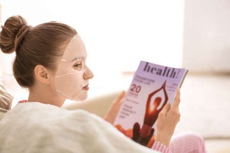 Young woman with sheet facial mask reading magazine at homeの写真素材
