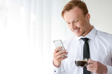 Handsome businessman with phone drinking coffee near window