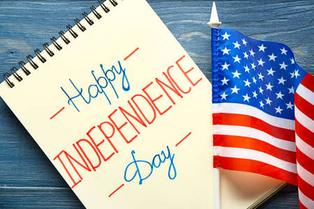 Notebook with text HAPPY INDEPENDENCE DAY and USA flag on color wooden background