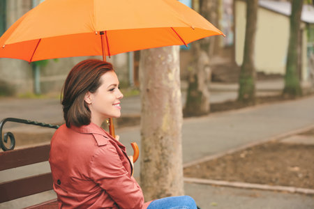 Beautiful young woman with umbrella sitting on bench outdoorsの写真素材