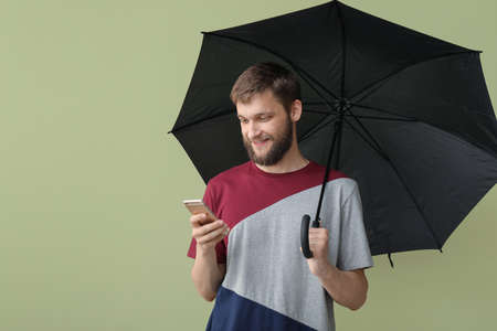 Handsome man with umbrella and mobile phone on color backgroundの写真素材
