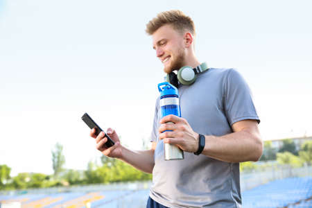 Sporty young man with mobile phone and bottle of water at the stadiumの写真素材