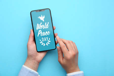 Female hands holding mobile phone with text WORLD PEACE DAY on screen against color background
