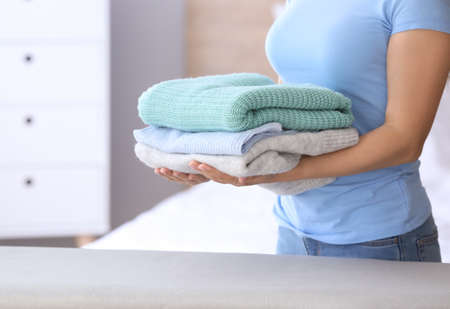 Young woman with stack of clean clothes standing near ironing board at home