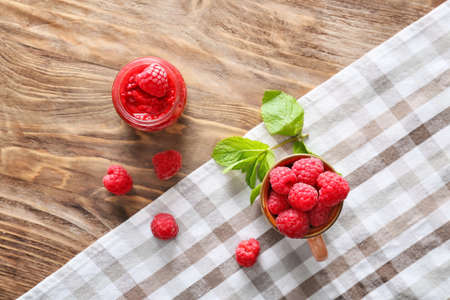 Cup with fresh raspberries and jam in jar on wooden table