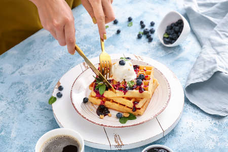 Woman eating tasty waffles with ice cream and blueberries at table