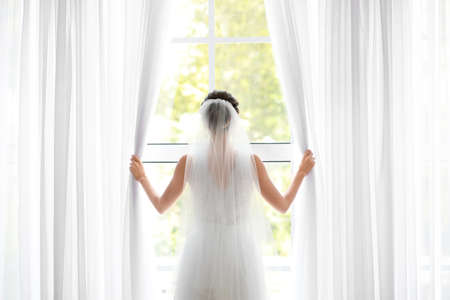 Beautiful young bride opening curtains on window before wedding ceremony at home, back viewの写真素材
