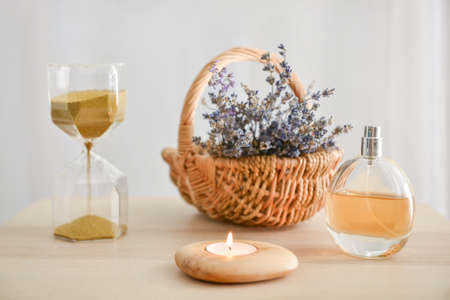 Burning candle with bottle of perfume, basket with lavender and hourglasses on table