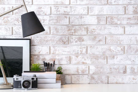 Comfortable workplace with lamp, photo camera, books and decor near brick wall