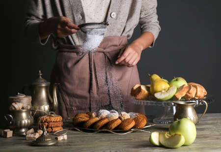Woman sprinkling sugar powder onto tasty cookies on table