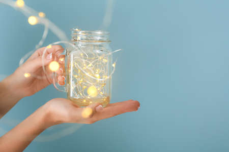 Female hands with glowing garland in jar on color background