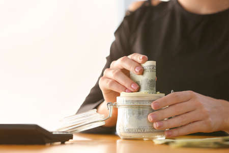 Woman putting dollar banknotes into jar on table