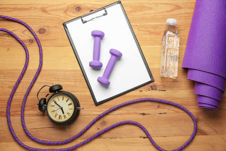 Dumbbells with clipboard, alarm clock, yoga mat, skipping rope and bottle of water on wooden backgroundの写真素材