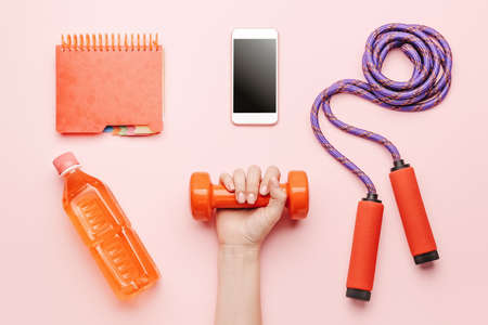 Female hand with dumbbell, bottle of water, mobile phone, notebook and jumping rope on color backgroundの写真素材
