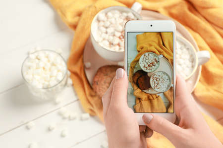 Female food photographer with mobile phone taking picture of hot chocolate with marshmallows
