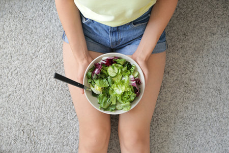 Woman with healthy vegetable salad in bowl sitting on floorの写真素材