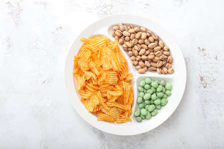 Plate with tasty potato chips, pistachios and peanuts on light background