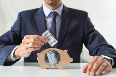 Male bank manager putting money into piggy bank at table in office, closeup