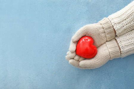 Female hands in warm gloves holding red heart on color background