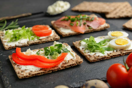 Crispbread with tasty cream cheese, salmon, eggs and vegetables on dark background, closeup