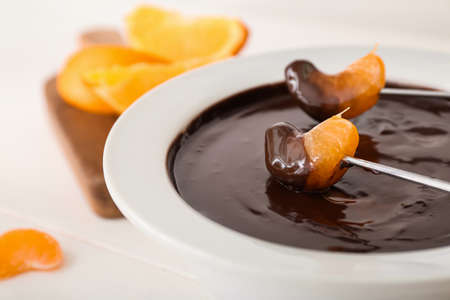 Dipping of tasty tangerine into bowl with chocolate fondue on table, closeupの写真素材