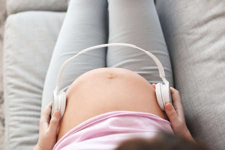 Beautiful pregnant woman putting headphones on her belly while sitting on sofaの写真素材