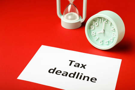 Paper sheet with text TAX DEADLINE, clock and hourglass on color backgroundの写真素材