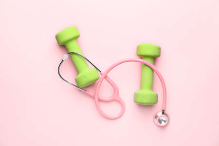 Stethoscope and dumbbells on color background