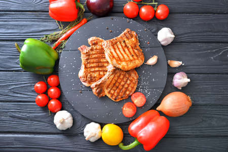 Board with cooked pork steaks and fresh vegetables on tableの写真素材