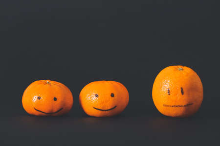 One citrus fruit with drawn neutral face among happy ones on dark background. Concept of depressionの写真素材