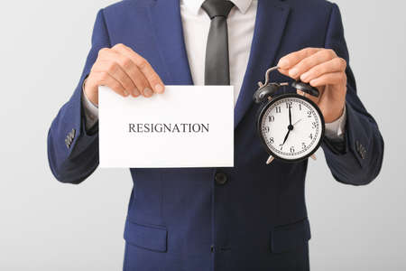Fired worker with alarm clock and paper with word RESIGNATION on light background