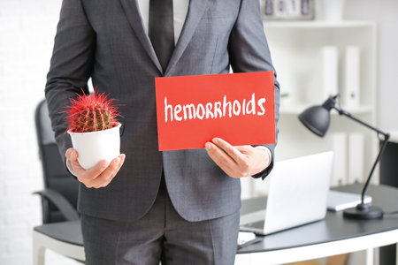 Young businessman holding cactus and paper with text HEMORRHOIDS in officeの写真素材