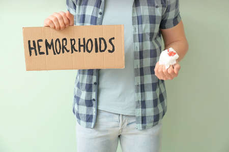 Young man holding paper with text HEMORRHOIDS and toilet paper in blood on color backgroundの写真素材