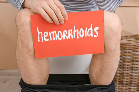 Young man with hemorrhoids sitting on toilet bowl in restroomの写真素材