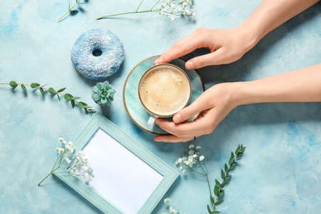Female hands with cup of coffee, blank photo frame, donut and flowers on color backgroundの写真素材
