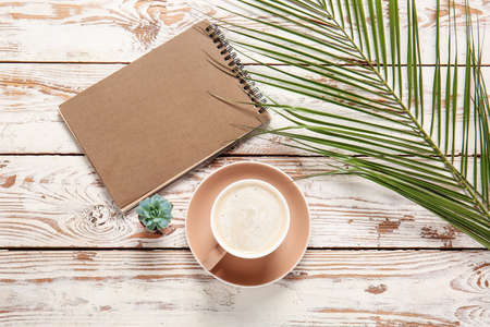 Cup of coffee, notebook and tropical leaf on light wooden backgroundの写真素材