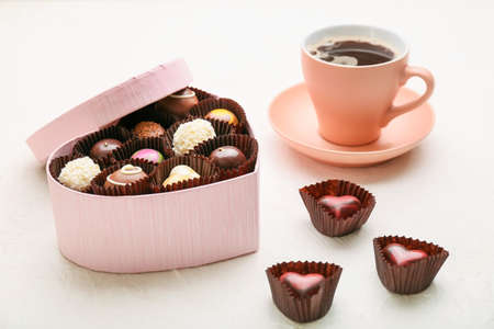 Heart-shaped box with delicious candies and cup of coffee on white background