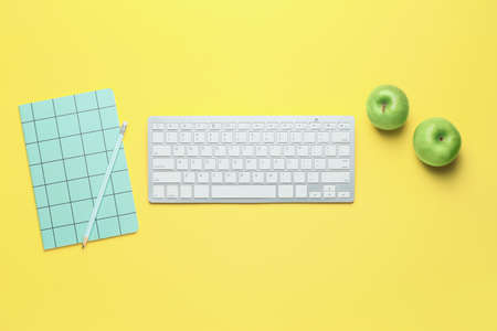 Computer keyboard with notebook and apples on color background