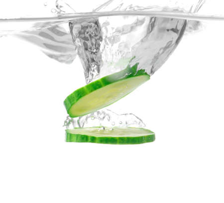 Falling of fresh cucumber slices into water against white backgroundの写真素材