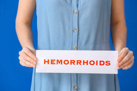 Young woman holding paper with text HEMORRHOIDS on color backgroundの写真素材