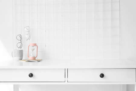 Cup, hourglass and eyeglasses at workplace near white wall