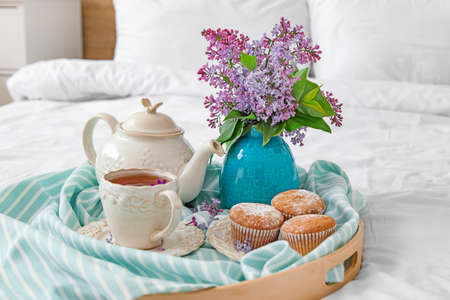Vase with beautiful lilac flowers, tea and muffins on tray in bedroomの写真素材